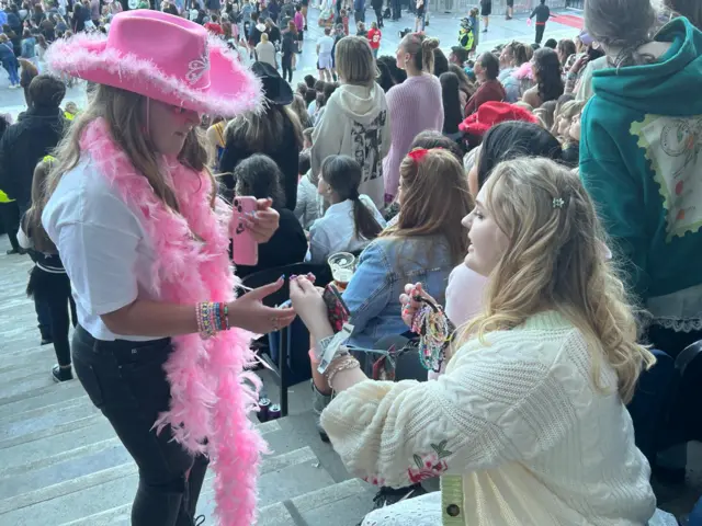 Two girls exchange friendship bracelets in a stadium, one wearing a pink cowboy hat and matching feather boa