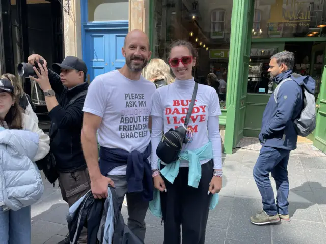 A man wearing a white shirt that reads 'Please ask my daughter about her friendship bracelets' and a woman wearing a white turtleneck which reads 'But daddy I love him' standing on a crowded Edinburgh street outside a shop