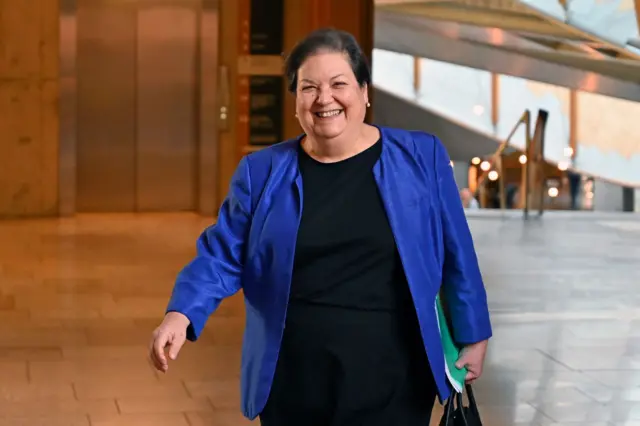 Dame Jackie Baillie walking in the atrium of the Scottish parliament, smiling and wearing a bright blue jacket and black dress and carrying a handbag and folder