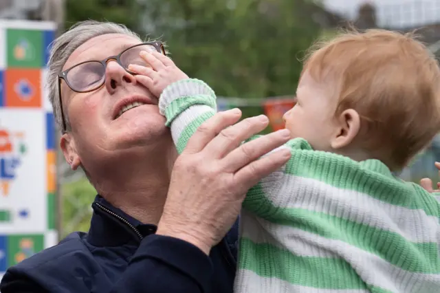 Starmer holds a small child you reaches up towards his face