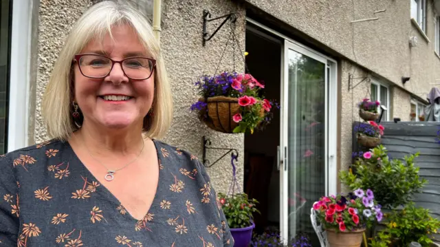 Image of a lady. She is blonde, wearing glasses and smiling at the camera. She is stood outside a house.