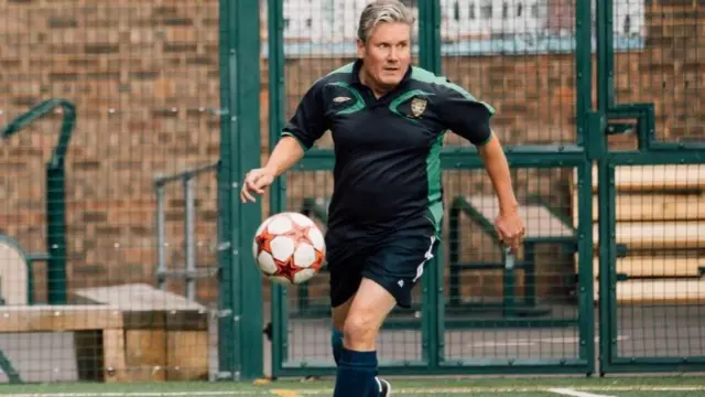 Keir Starmer wearing football kit and about to kick football in caged astro turf pitch