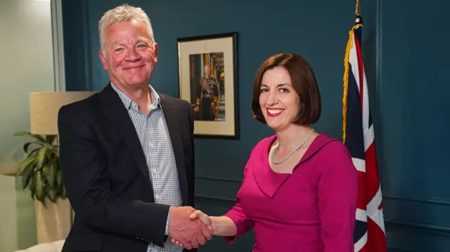 Sir Kevan Collins, who has short grey hair and wears a dark jacket and blue chequered shirt, smiles at the camera as he shakes hands with Education Secretary Bridget Phillipson. She has short, straight, brown hair, and wears a bright pink dress. They are standing in an office in front of a Union Jack flag and a framed portait of King Charles