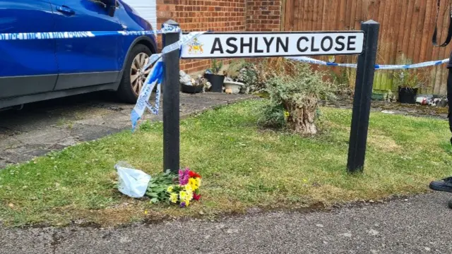 A bouquet of flowers is laid on the grass in front of an 'Ashlyn Close' road sign. Police tape is held up by the sign