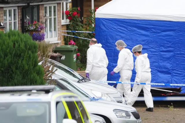 Three forensic officers walk past a blue tent heading towards a house