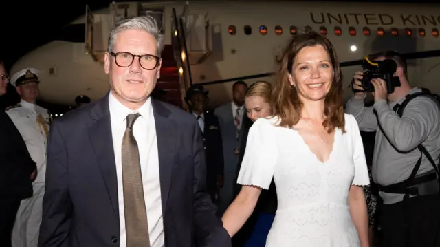 Keir Starmer and his wife Victoria disembark from their plane in the dark with photograph behind them taking picture