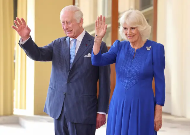 King Charles wearing a blue suit and tie while Queen Camilla is wearing a blue dress.