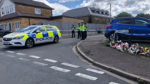 Ashlyn Close with bunches of flowers on the ground to the right, and uniformed police officers stand behind a police car and police cordon