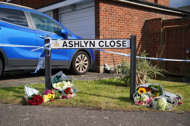 Bouquets of flowers left under an "Ashlyn Close" street sign, with a blue car behind the sign.