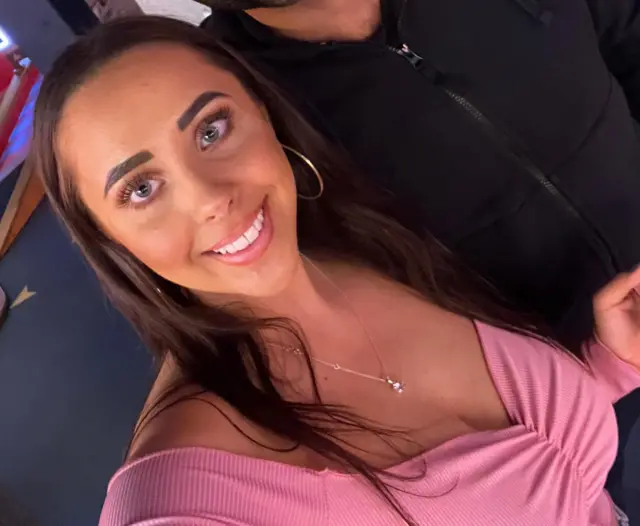 A smiling Hannah Hunt looking directly at the camera, with long dark hair and wearing a pink top