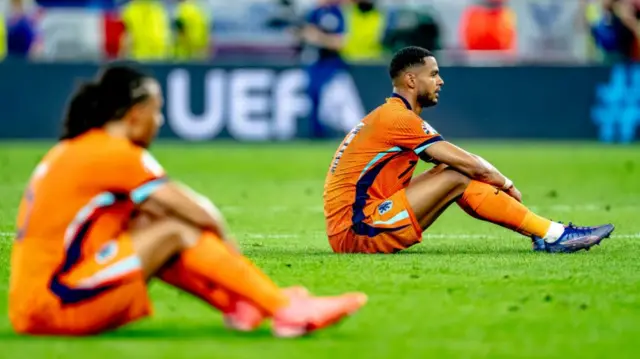 Nathan Ake and Cody Gakpo sat on the turf looking disappointed after the Netherlands lose to England at Euro 2024