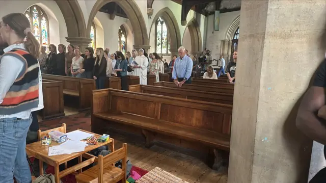 Attendees of the service stand in pews inside of St James's Church in Bushey. Some have their heads bowed.