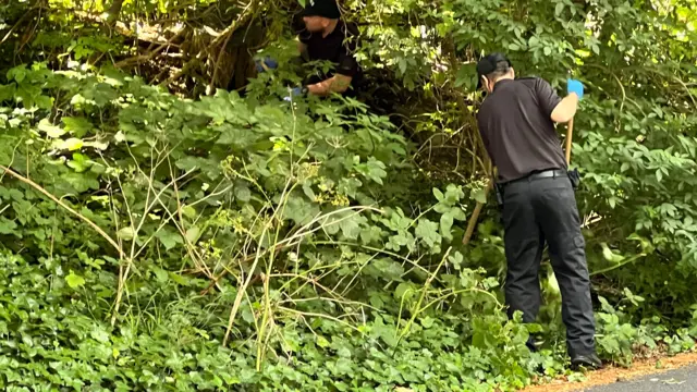 Two officers wearing black clothing and hats searching an area of woodland.