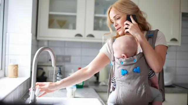 A woman pours a glass of water while carrying her baby in a sling