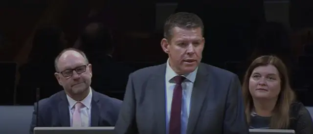 Rhun ap Iorwerth standing in the Senedd wearing a suit