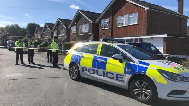 Police cordon outside two-storey brick and timber house. Police car in foreground. Three police officers behind police tape.