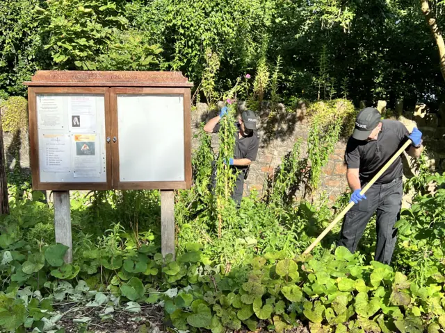 Two officers searching an area of Leigh Woods. A wooden sign can be seen to the left of the frame, and two men searching