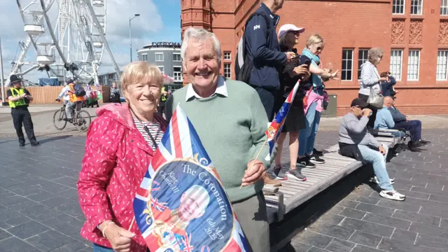 A man and woman holding union jack flags. The woman is wearing a pink raincoat while the man is wearing a light green jumper with a white shirt underneath. They are both smiling at the camera.