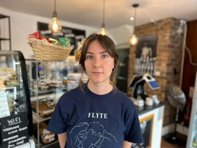 Joely Rendle. She has dark brown hair which is tied up. She is in a cafe, wearing a black t-shirt. She is looking directly at the camera and smiling.