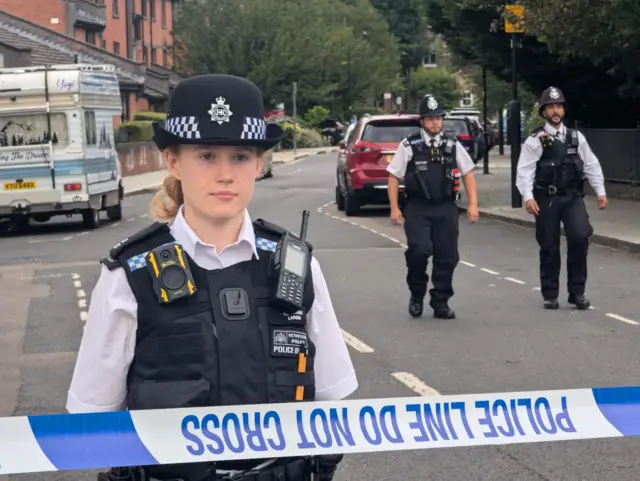 A female police officer standing behind police tape with two male officers behind her