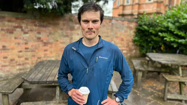 Dan Summers. He has dark brown hair and a small amount of facial hair. He is wearing a blue jacket and carrying a brown takeaway coffee cup.