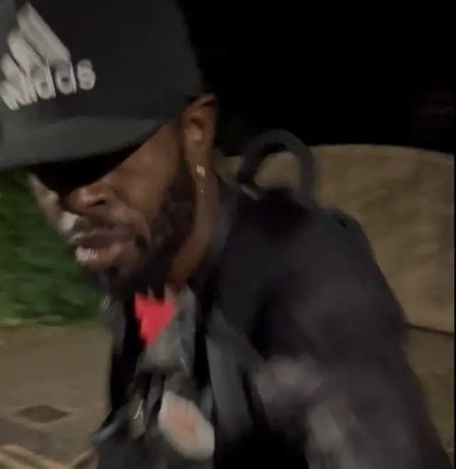 A photo of a black man wearing all black and a black Adidas cap
