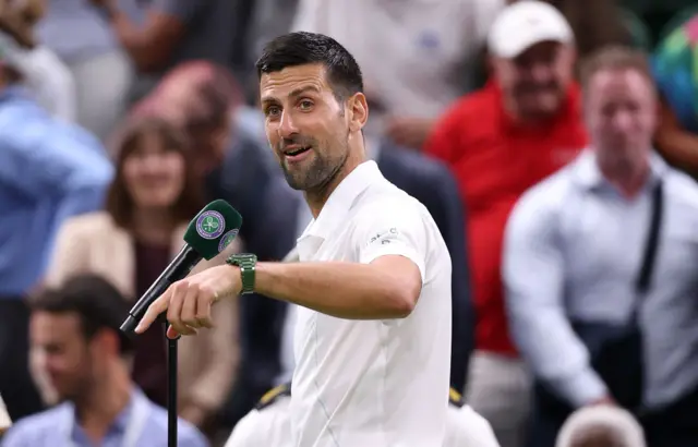 Novak Djokovic stand in front of a microphone and points to the floor