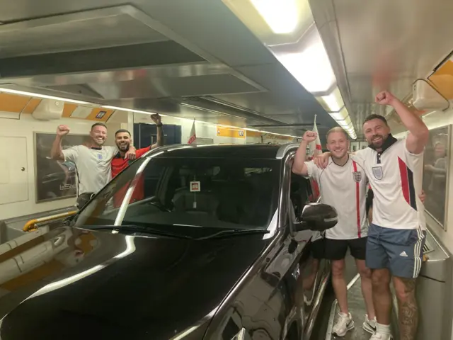 Four men, dressed in white England football jerseys, pose outside a black car inside the Eurotunnel