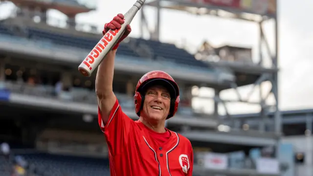 Representative Steve Scalise wears a red baseball uniform and holds up a baseball bat