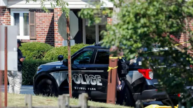 A police car sits outside a brick home with authorities standing outside and police tape blocking the premises