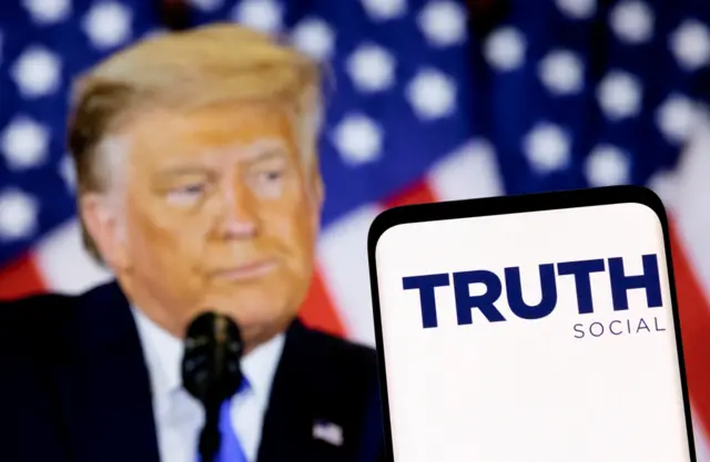 The Truth social network logo is seen on a smartphone in front of a display of former US President Donald Trump