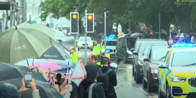 Convoy making its way past people waving flags while holding umbrellas