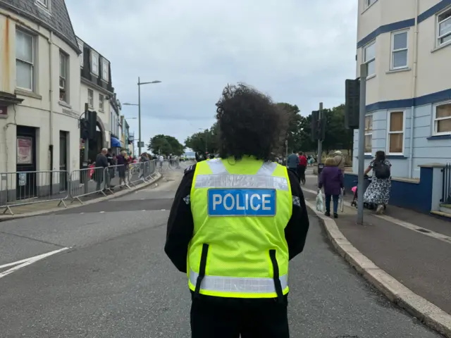 Police officer stood in road with barriers at the side of the road and people walking past