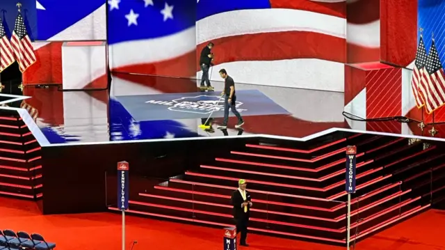 Two people mop a stage with red steps leading up to it and an American flag as its backdrop