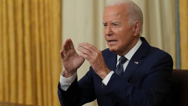 Biden at desk in front of gold curtains, with hands raised