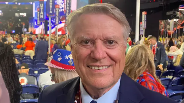 Greg Simpson stands next to rows of seats at the convention, smiling at the camera