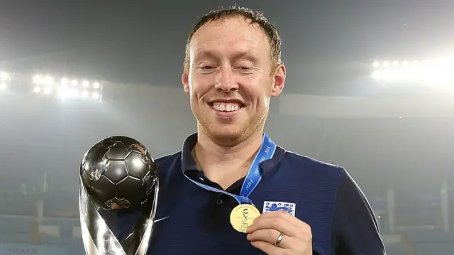 Steve Cooper, wearing a dart blue shirt with England's Three Lions badge on, holds the silver Under-17 World Cup trophy in his right hand and lifts up his gold winners medal to the camera with his left hand