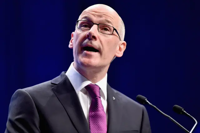 A man with a black suit and purple tie speaking in front of a microphone.