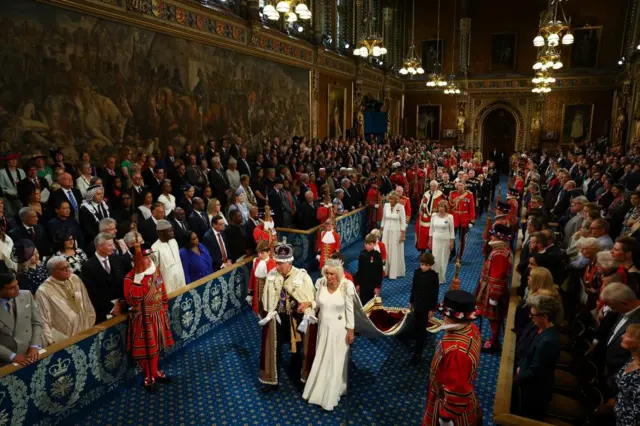 King Charles III, wearing the Imperial State Crown and the Robe of State, and Queen Camilla, wearing the George IV State Diadem, walk through the Royal Gallery during the State Opening of Parliament