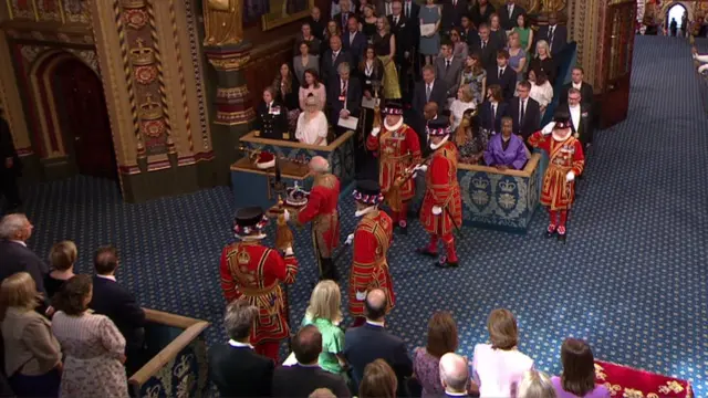 Crown carried by man in bright red regalia as people stand either side looking on