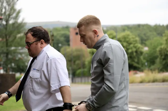 Cameron Jones wearing a grey jacket, walking while handcuffed to a security guard in a white shirt and black tie