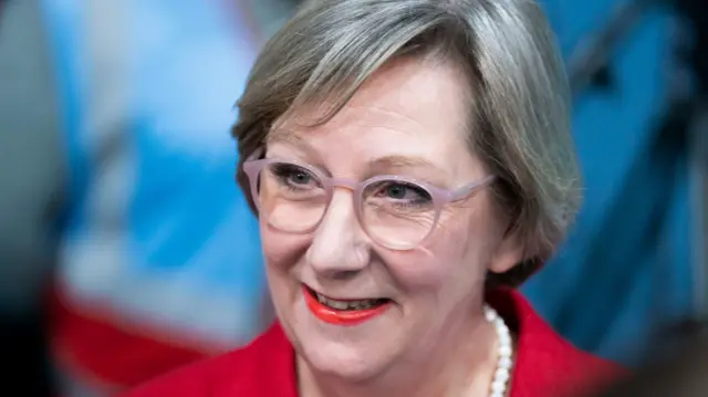 A headshot of Samantha Dixon .She's wearing pale pink glasses, a red jacket, a pearl necklace and red lipstick.