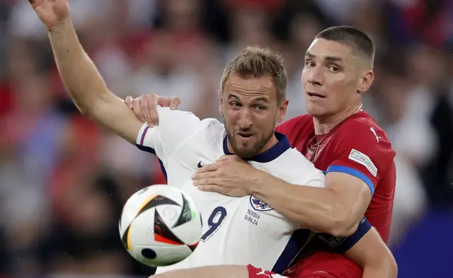 Nikola Milenkovic, wearing Serbia's red shirt, grapples with England captain Harry Kane with his left arm over his chest and right arm holding his shoulder.