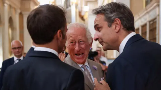 King Chalres laughing looking at the president of France (with his back turned), the prime minister of Greece is to the rigth.