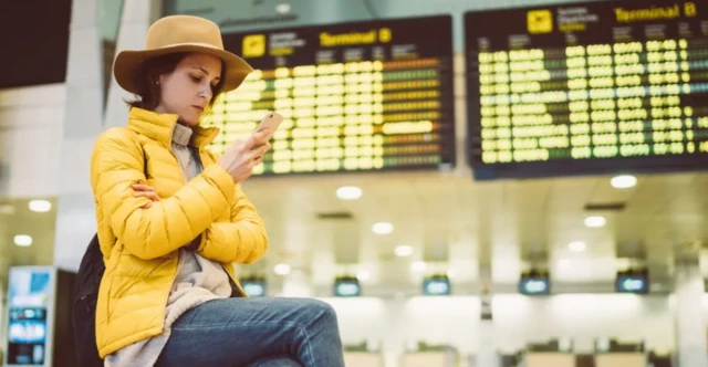 A woman in a yellow coat looks at her phone by an airport departure board.