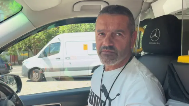 Head shot of taxi driver in a London taxi looking out of the left side window, there is a white van in the background