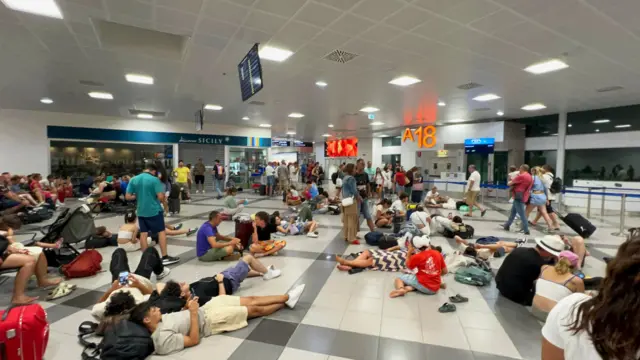 People lie on the floor in an airport, resting their heads on bags