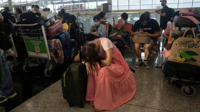 Woman in pink head sits laying her head on a suitcase while people around sit on chairs next to bags and suitcases