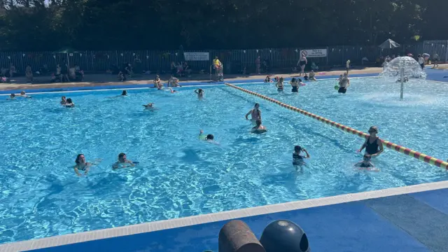 A blue lido pool in the sunshine. People are swimming and playing in the water