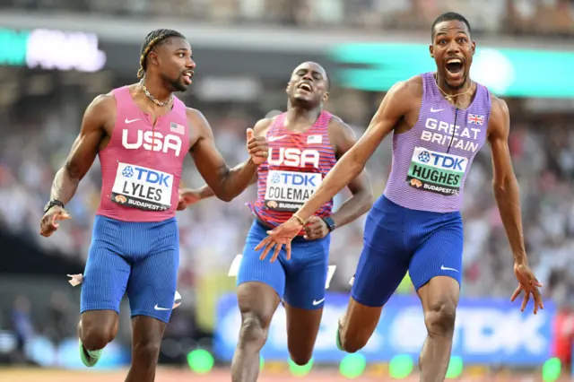 Zharnel Hughes holds out his arms and smiles as he runs next to Noah Lyles and Christian Coleman